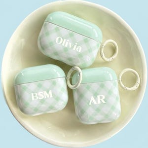 May include: Three mint green and white gingham patterned AirPod cases with personalized initials and names. Each case has a matching mint green top and a small metal ring for attachment. The cases are displayed in a light yellow bowl.