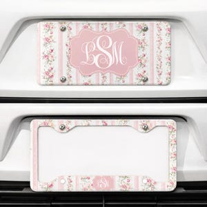 May include: A car license plate with a pink and white floral design. The plate features a pink central emblem with the letters "LSM" in white script. The background has vertical pink stripes and floral accents.