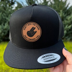 May include: Black trucker hat with a curved brim and mesh back. The hat features a circular patch with the text "DON'T BITE THE HAND THAT FINGERS YOU" and a hand gesture. The hat has a snapback closure.