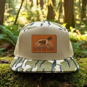 May include: A camouflage trucker hat with a brown leather patch featuring a dog illustration and the text "GET TO THE POINT." The hat has a tan mesh back and a curved brim with a green and blue camouflage pattern.