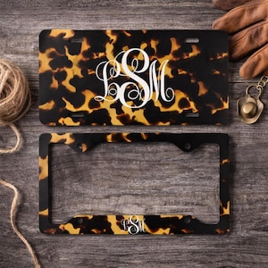 May include: Tortoiseshell-patterned license plate and frame with a white monogram. The license plate has a black, brown, and yellow pattern. The frame is black with a matching tortoiseshell pattern. A decorative item for a vehicle.