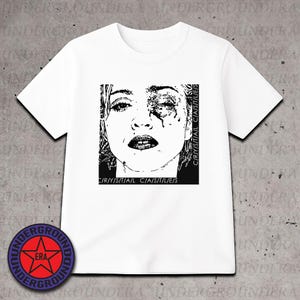 May include: White t-shirt featuring a black and white graphic of a woman's face with the text "CRYSTAL CASTLES" and "C/R/Y/S/T/A/L C/A/S/T/L/E/S". The design is a bold, artistic statement. The shirt has a classic crew neck and short sleeves.