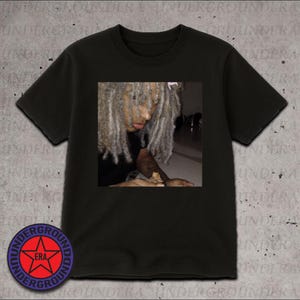 May include: Black t-shirt featuring a square image of a person with gray dreadlocks. The person is looking down, with a blurred background. The shirt has a round logo on the bottom left that says "UNDERGROUND ERA".