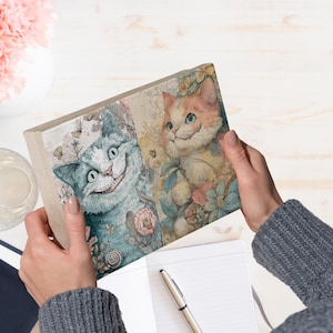 May include: A rectangular journal featuring two smiling cat illustrations. One cat is blue, the other is orange, both surrounded by floral and botanical elements. The journal is held open, revealing blank pages.