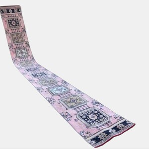 May include: A long, rectangular pink runner rug with a repeating pattern of square medallions. The medallions feature intricate designs in shades of blue, beige, and black. The rug has a dark border and a red edge, suitable for a hallway or entryway.