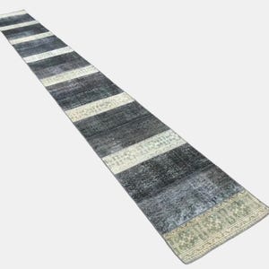 May include: A long, rectangular runner rug with alternating stripes of dark grey and off-white. The rug has a distressed, vintage appearance, with a patterned border in shades of green and beige. Suitable for hallways or entryways.
