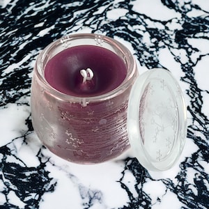 Könnte beinhalten: Ein kleiner, klarer Kerzenhalter aus Glas mit einer tiefvioletten Kerze im Inneren. Die Kerze hat einen weißen Docht. Der transparente Deckel liegt daneben. Der Kerzenhalter und der Deckel haben ein dekoratives Muster. Der Hintergrund ist ein schwarz-weißer Marmor.