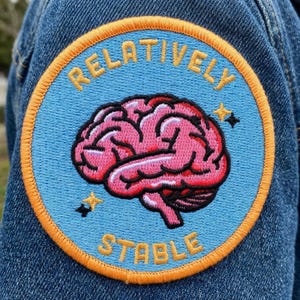 May include: Embroidered patch with a light blue background and an orange border. The patch features a pink brain graphic with the words "RELATIVELY" and "STABLE" in yellow lettering. Two small star graphics are also included.