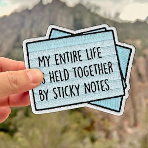May include: A light blue patch with the text "MY ENTIRE LIFE HELD TOGETHER BY STICKY NOTES" in black. The patch is in the shape of two overlapping sticky notes with a black border. The background is a blurred mountain scene.