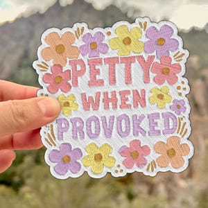 May include: Embroidered sticker with the text "PETTY WHEN PROVOKED" in pink and purple. The text is surrounded by colorful embroidered flowers in shades of pink, yellow, and purple. The sticker is held by a hand.