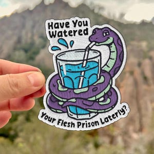 May include: Embroidered patch featuring a purple snake coiled around a glass of blue liquid with a straw. The patch has the text "Have You Watered Your Flesh Prison Lately?" in white. The patch is held by a hand.