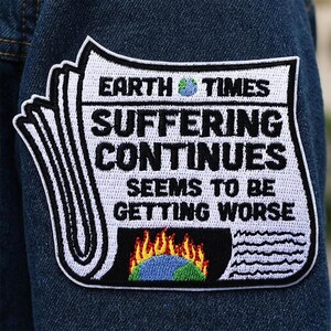May include: Embroidered patch with the text "EARTH TIMES SUFFERING CONTINUES SEEMS TO BE GETTING WORSE" on a white background. The patch features a globe and flames, and is sewn onto a denim jacket.