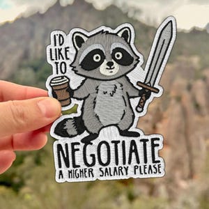 May include: Embroidered patch featuring a cartoon raccoon holding a coffee cup and a sword. The patch reads "I'D LIKE TO NEGOTIATE A HIGHER SALARY PLEASE" in black text with a white border. The raccoon is gray, black, and white.