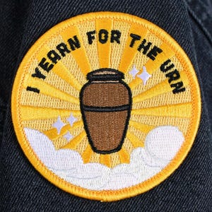 May include: Embroidered patch with a yellow border and a brown urn design. The patch features the text "I YEARN FOR THE URN" in a semi-circle around the urn. The background has a sunburst design with white clouds and stars.