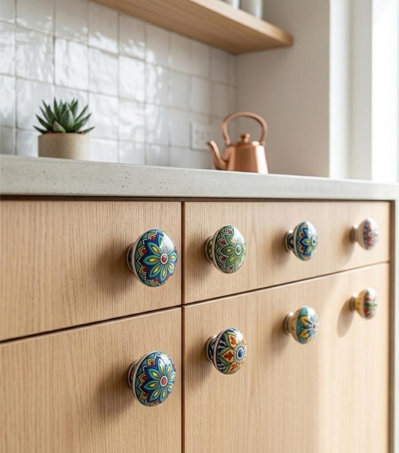 以下が含まれることがあります： 明るい木製のキャビネットのクローズアップ。カラフルなセラミックノブが付いています。ノブは、青、緑、黄色、赤の色合いの複雑な花柄のデザインが特徴です。銅製のティーポットと小さな鉢植えの多肉植物がカウンタートップにあります。