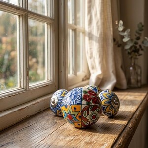 以下が含まれることがあります： 木製の窓枠に、カラフルな花柄と幾何学模様の装飾的なセラミックキャビネットノブが4つ置かれています。ノブは青、黄、赤、白のデザインで、家の装飾に芸術的な雰囲気を添えています。