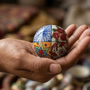 以下が含まれることがあります： 手描きの装飾的なセラミック球体で、セグメント化されたデザインです。各セクションには、青い花柄、赤いバラ、スターバースト、花柄など、ユニークなパターンがあります。球体は開いた手に持たれており、その複雑なディテールが示されています。