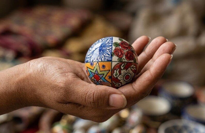 以下が含まれることがあります： 手描きの装飾的なセラミック球体で、セグメント化されたデザインです。各セクションには、青い花柄、赤いバラ、スターバースト、花柄など、ユニークなパターンがあります。球体は開いた手に持たれており、その複雑なディテールが示されています。