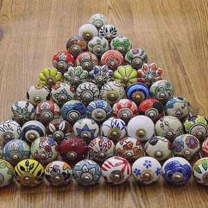 以下が含まれることがあります： 三角形に配置された、装飾的なセラミックキャビネットノブのコレクション。ノブは、真鍮色の金具を備えた、さまざまなカラフルな花柄と幾何学模様が特徴です。ノブは木製の表面に表示されています。