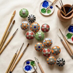 以下が含まれることがあります： 様々な花柄や幾何学模様が施された、カラフルなセラミック製のキャビネットノブのコレクション。ノブは、絵の具パレット、筆、小さなテラコッタのポットと一緒に配置されており、芸術的なテーマを示唆しています。ノブはオレンジ、緑、青、赤の混合色です。