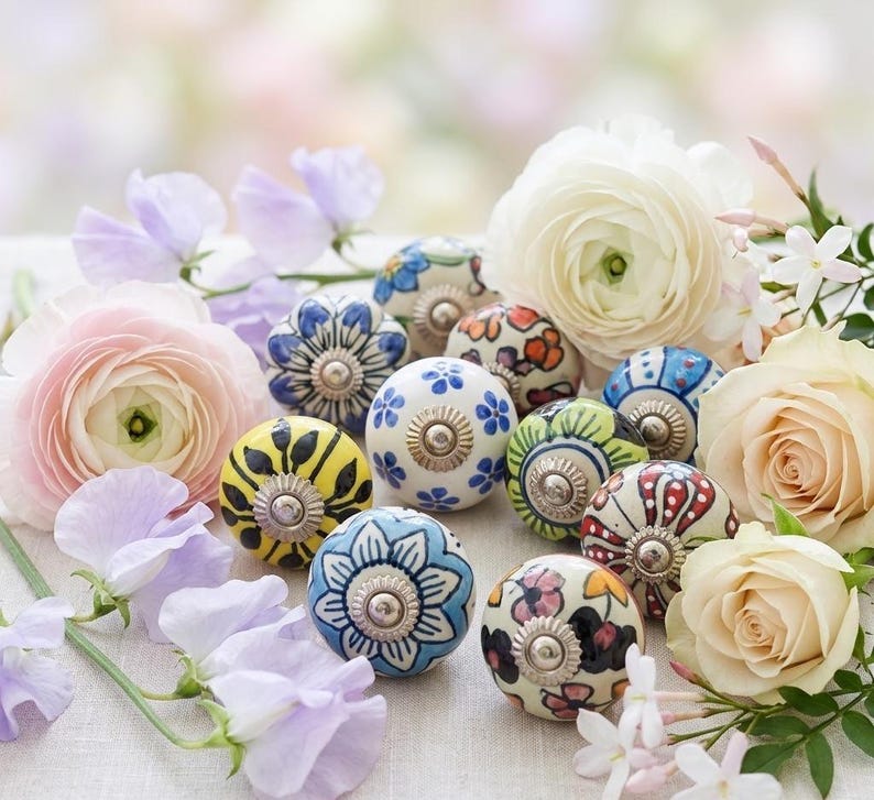 以下が含まれることがあります： 青、黄色、赤など、さまざまな色の花柄が施された装飾的なセラミックキャビネットノブのコレクション。パステルカラーの花々の間にノブが配置されています。