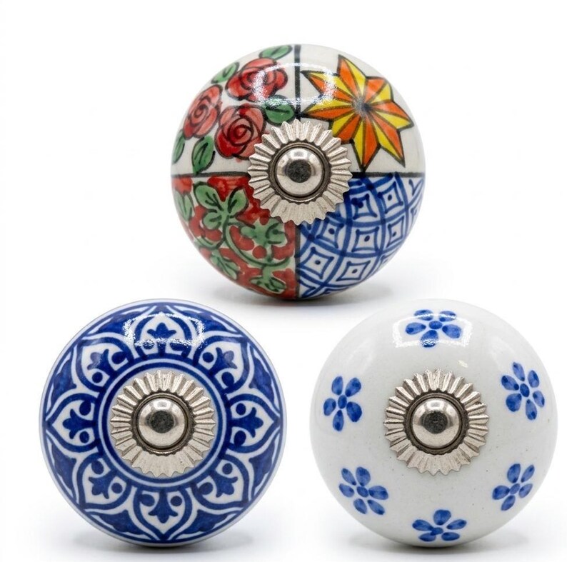 以下が含まれることがあります： 3つの装飾的なセラミックキャビネットノブ。1つのノブは、赤いバラ、黄色の星、青い幾何学模様のフローラルデザインが特徴です。他の2つのノブは、青と白の花柄です。各ノブには、銀色の金属ベースが付いています。