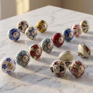 以下が含まれることがあります： 様々な色と模様の装飾的なセラミックキャビネットノブのコレクション。デザインには、花柄、幾何学模様、抽象的なモチーフが含まれます。各ノブは金属製のベースとユニークな塗装デザインを備えています。