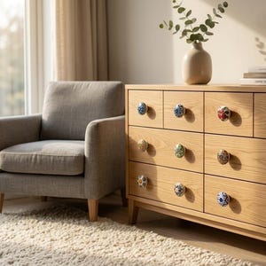 以下が含まれることがあります： 9つの引き出しが付いた明るい木製のドレッサー。各引き出しには、ユニークでカラフルなセラミックノブが付いています。左側には灰色のアームチェアがあり、ドレッサーの上には緑のある花瓶が置かれています。部屋は自然光で満たされています。