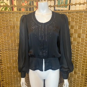 May include: A black satin blouse with long sleeves and a gathered waist. The blouse features a round neckline and a beaded design on the front.