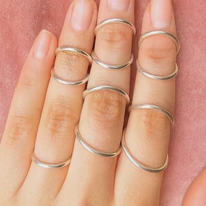 Può includere: Primo piano di una mano con più dita adornate da anelli midi in argento. Gli anelli sono semplici fasce curve, indossate sulle nocche medie e superiori. Lo sfondo è rosa tenue.