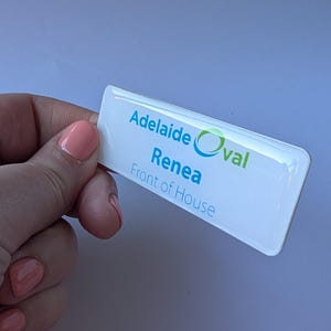 May include: A white rectangular name badge with rounded corners. The badge displays the text "Adelaide Oval", "Renea", and "Front of House" in blue and green. The badge is held by a person.