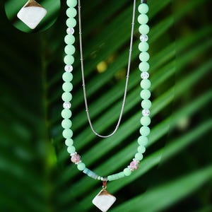 Puede incluir: Un collar de varias capas con una cadena plateada y una hebra de cuentas verde menta. El collar incluye un colgante con una piedra cuadrada blanca y ribete dorado. También hay cuentas adicionales en rosa y transparente.