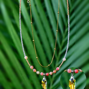 Op de afbeelding: Een gelaagde ketting met een goudkleurige ketting met kleine kraaltjes en een tweede streng van lichtroze en witte zaadkraaltjes. Het onderste gedeelte bevat roze en oranje crackle-kralen met een gele kristallen hanger in de vorm van een vleugel.