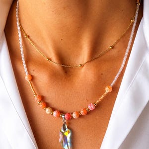 Op de afbeelding: Een gelaagde kettingenset bestaande uit een fijne gouden ketting met kleine, gelijkmatig verdeelde kraaltjes en een tweede streng met transparante, oranje en roze craquel&eacute; kralen met een centrale iriserende kristallen hanger in de vorm van een vleugel.