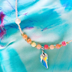 Op de afbeelding: Een kralenketting met een centrale iriserende, vleugelvormige hanger aan een zilverkleurig oogje. Het snoer bestaat uit kleine lichtroze zaadkralen, een enkele grote lichtroze kraal en een reeks gebarsten oranje, gele en roze kralen.