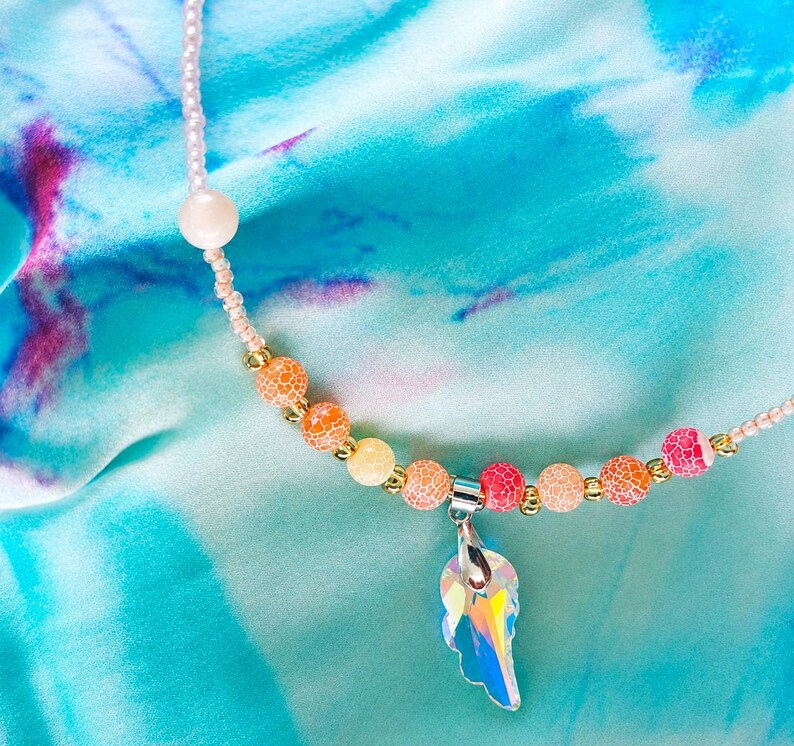 Op de afbeelding: Een kralenketting met een centrale iriserende, vleugelvormige hanger aan een zilverkleurig oogje. Het snoer bestaat uit kleine lichtroze zaadkralen, een enkele grote lichtroze kraal en een reeks gebarsten oranje, gele en roze kralen.