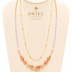 Op de afbeelding: Een gelaagde ketting met een fijne gouden ketting en een streng kleine lavendelkleurige, perzikkleurige en koraalkleurige kralen. De ketting heeft een centrale gefacetteerde barnsteenkleurige kristallen hanger en kleine gouden metalen accenten.