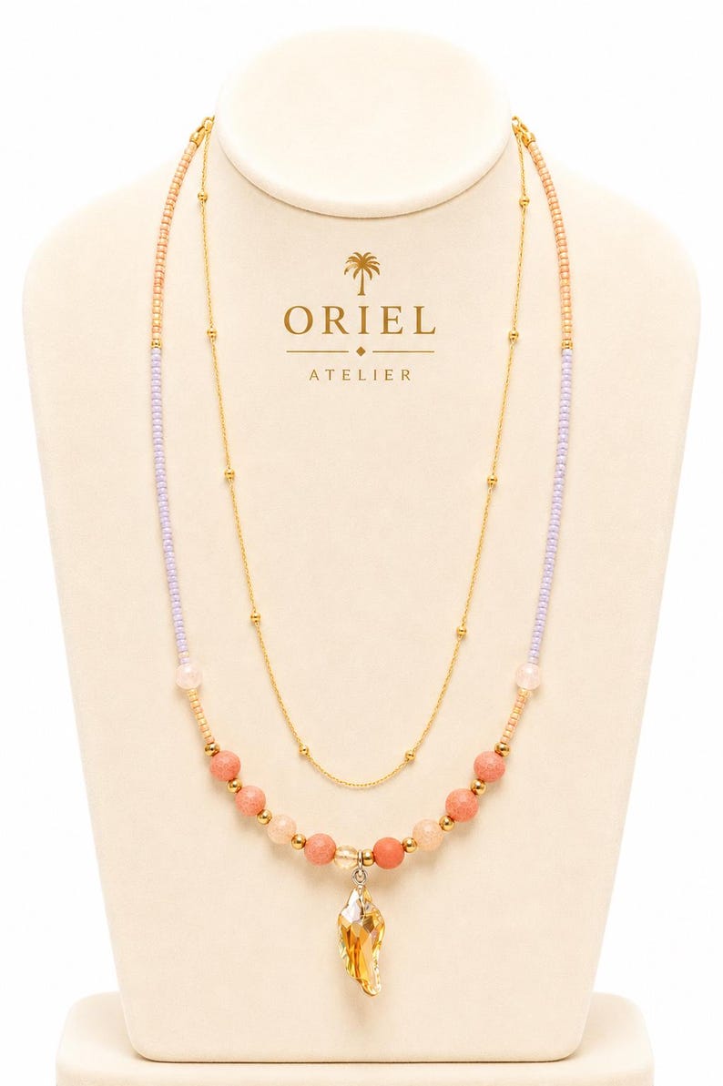 Op de afbeelding: Een gelaagde ketting met een fijne gouden ketting en een streng kleine lavendelkleurige, perzikkleurige en koraalkleurige kralen. De ketting heeft een centrale gefacetteerde barnsteenkleurige kristallen hanger en kleine gouden metalen accenten.
