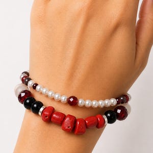 May include: A two-strand bracelet set featuring one strand of small white pearls interspersed with faceted dark red beads. The second strand consists of larger, irregular red coral-like pieces, smooth black beads, and faceted dark red beads with silver spacers.