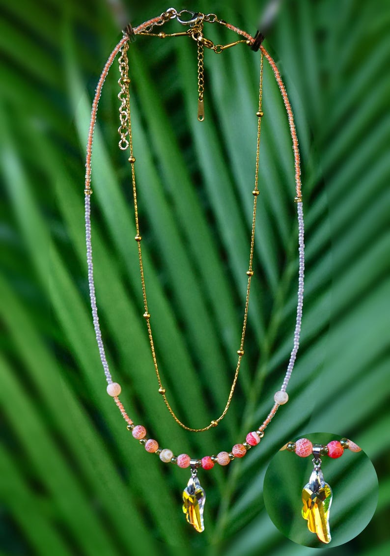 Op de afbeelding: Een gelaagde ketting met een goudkleurige ketting met kleine kraaltjes en een tweede streng van lichtroze en witte zaadkraaltjes. Het onderste gedeelte bevat roze en oranje crackle-kralen met een gele kristallen hanger in de vorm van een vleugel.