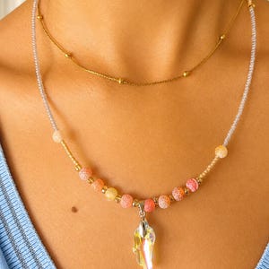 Op de afbeelding: Een gelaagde kettingenset bestaande uit een dunne gouden ketting met kleine gouden kralen en een tweede streng van transparante zaadkralen, afgewisseld met oranje en roze craquel&eacute; agaatkralen, afgewerkt met een heldere iriserende kristallen hanger.