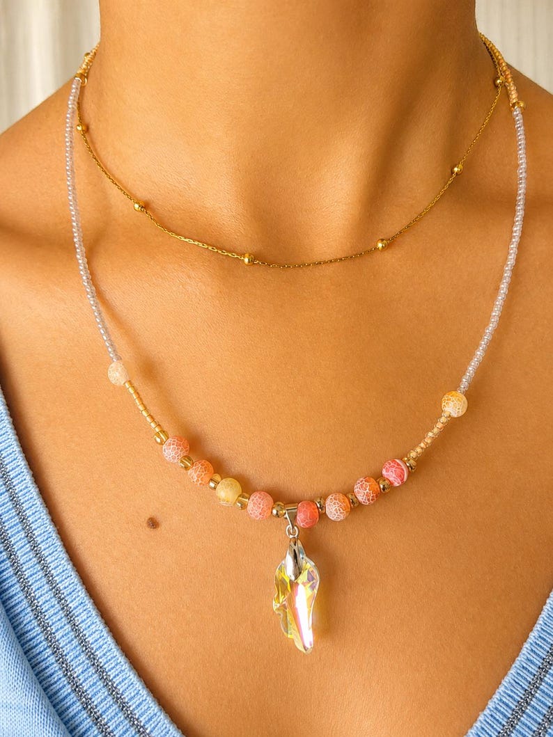 Op de afbeelding: Een gelaagde kettingenset bestaande uit een dunne gouden ketting met kleine gouden kralen en een tweede streng van transparante zaadkralen, afgewisseld met oranje en roze craquel&eacute; agaatkralen, afgewerkt met een heldere iriserende kristallen hanger.