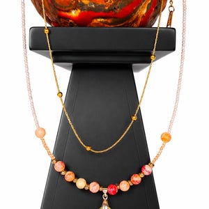 Op de afbeelding: Een meerrijige ketting met een fijne gouden ketting en een rij lichtroze kraaltjes. De ketting is versierd met ronde, gebarsten oranje, rode en gele kralen en een centrale geel-oranje druppelvormige kristallen hanger.