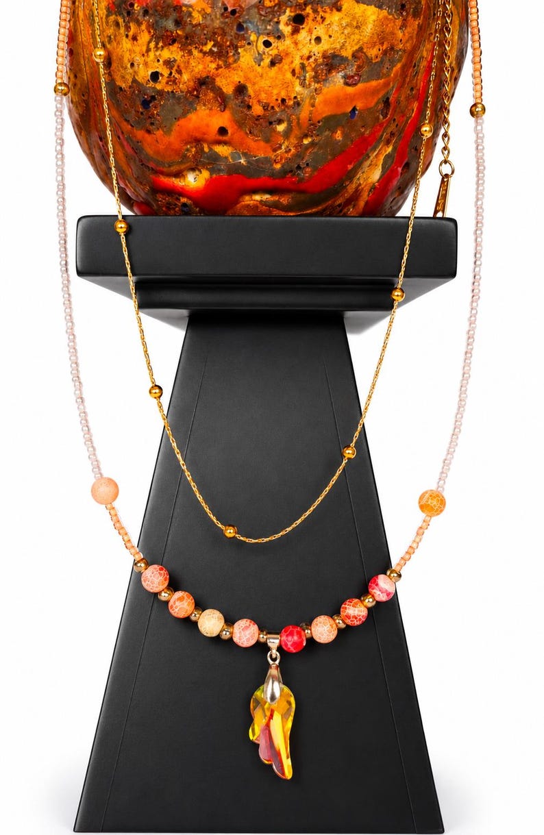 Op de afbeelding: Een meerrijige ketting met een fijne gouden ketting en een rij lichtroze kraaltjes. De ketting is versierd met ronde, gebarsten oranje, rode en gele kralen en een centrale geel-oranje druppelvormige kristallen hanger.