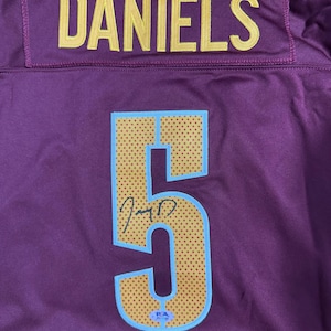 Pode incluir: Uma camisola desportiva bordeaux com o nome DANIELS em letras amarelas na parte superior das costas. Abaixo, um grande número 5 em dourado com contorno azul claro e textura pontilhada, com uma assinatura preta e um autocolante PSA.
