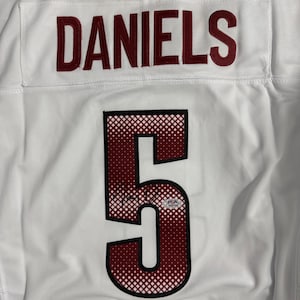 Pode incluir: Uma camisola desportiva branca com o nome DANIELS em letras vermelho-escuras acima do número 5. O número 5 tem um padrão de grelha vermelho-escuro e branco com um contorno preto. Um pequeno logótipo circular Commanders está no topo.