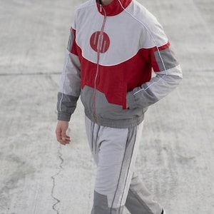 Könnte beinhalten: Ein grauer und roter Trainingsanzug mit einem kreisförmigen Logo auf der Brust. Die Jacke hat einen roten Kragen und einen Reißverschluss. Die Hose ist grau mit einem dunkleren grauen Einsatz an den Unterschenkeln. Schwarze Turnschuhe vervollständigen das Outfit.