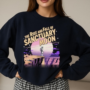 Puede incluir: Sudadera azul marino con un gráfico de un astronauta en un acantilado, con un gran luna rosa y blanca. El texto "THE RISE AND FALL OF SANCTUARY MOON" está impreso arriba. El diseño tiene una estética retro con temática espacial.