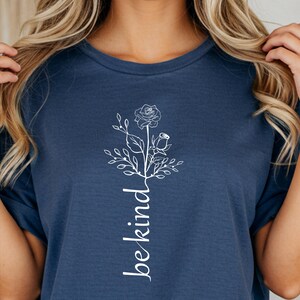 Könnte beinhalten: Marineblaues T-Shirt mit weißem Blumendesign und dem vertikalen Schriftzug "be kind". Die Grafik enthält eine Rose und andere florale Elemente. Das T-Shirt ist einfarbig und hat kurze Ärmel.