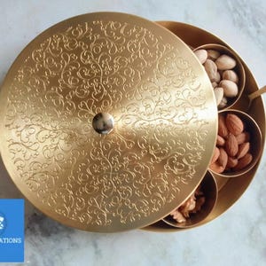 Könnte beinhalten: Eine goldfarbene Servierschale mit Deckel und aufwendigem floralem Muster. Die Schale ist in Fächer unterteilt, die mit verschiedenen Nüssen gefüllt sind. Ein kleiner Löffel liegt daneben. Das Logo "AMAYA CREATIONS" ist zu sehen.